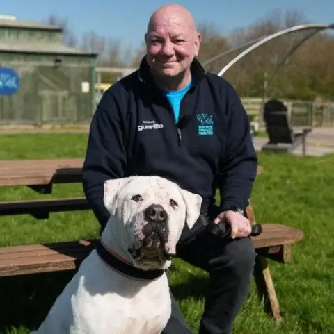 Matty sits on a bench with Yeti, a large white American bulldog by his feet. Matty is totally bald and clean shaven, while Yeti is a large dog with a large head.