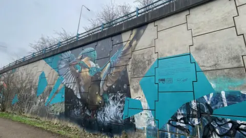 A colourful mural of a kingfisher is painted on a concrete wall. There are railings at the top and a street light.