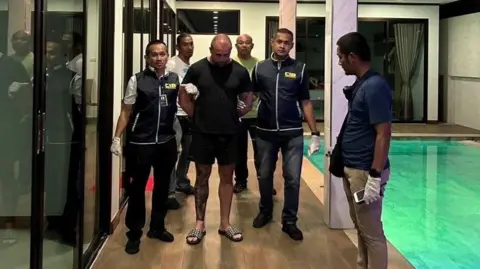 Essex Police Ransome being led away by several police officers after his arrest. They are holding his arms held behind his back. He is wearing the same black T-shirt as well as dark shorts and sliders. There is a swimming pool beside him.