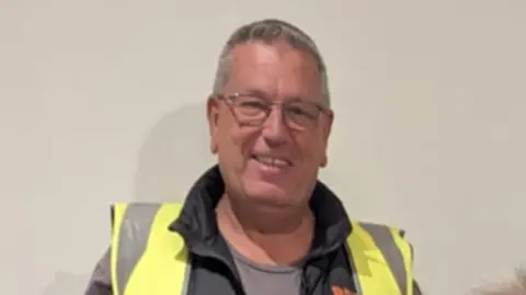 Band of Builders Tony Steel with short grey hair and glasses is standing against a grey wall. He is wearing a fluorescent hi-vis jacket.