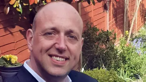 A man with very little hair, wearing a white shirt and a blue jacket and standing in front of a wooden fence in a garden smiles into the camera