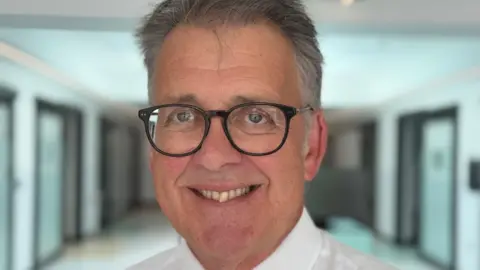 A man smiles broadly with black glasses and grey hair in a quiff