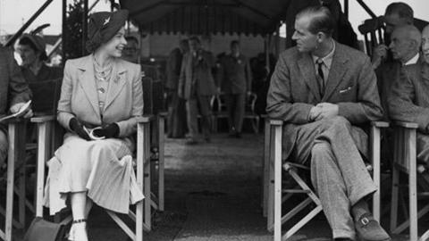Black and white image of Princess Elizabeth and Prince Philip in the foreground, prominently seated on two deck chairs amongst a seated crowd.  Elizabeth on the left is smiling across towards Philip on the right who is slightly more pensive looking.