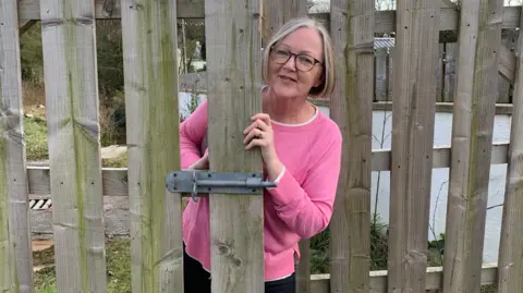 A woman in a pink jumper with glasses and short blonde hair looks out from behind a wooden gate