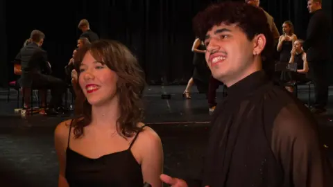 A young teenage girl and boy stand together smiling, talking off to the left of the camera, in a dark theatre hall/stage area