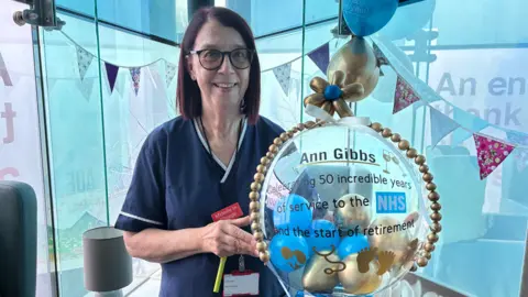 A woman with short, straight, brown hair and glasses holding a balloon with a message printed on it, stating: Ann Gibbs celebrating 50 incredible years of service to the NHS and the start of retirement.
She is wearing navy NHS staff uniform and in the background there are decorations and balloons. 
