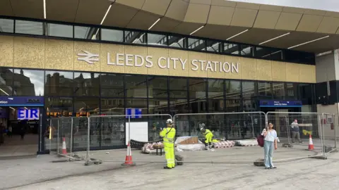 The front of Leeds railway station. There is a gold sign above a glass frontage, with the words "Leeds City Station" in white. There are safety barriers in front of the windows, and workers in hi-vis clothing can be seen. Some passengers are also standing around the entrance.
