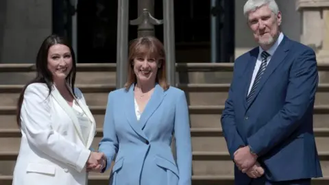 Reuters Emma Little Pengely, Ravel Reeves et John O'Dowd sont ensoleillés à l'extérieur de Stormont. Emma a de longs cheveux brun foncé et porte un dessus avec une veste blanche et un pantalon noir. Rachel a les cheveux bruns / rouges et porte un costume bleu clair et un haut blanc.