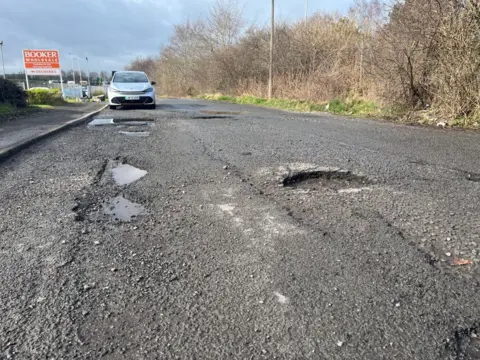 Several large potholes. A car is parked in the background and there is a sign for Bookers. 