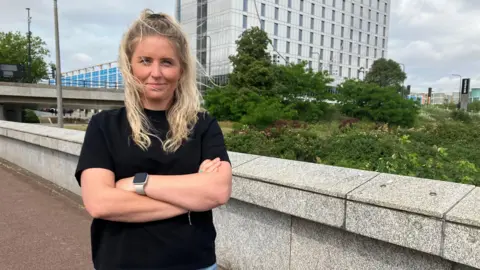 Amy Holmes/BBC A blonde-haired woman with crossed arms. She is standing on a path in Milton Keynes beside a stone wall, with greenery, an overpass, and a modern building in the background under a cloudy sky.