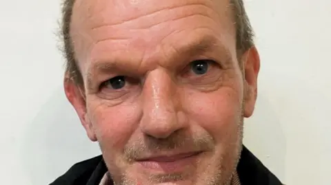 family handout A close-up of a Nick Starmer, who has light skin and short hair that is thinning at the top. His expression is neutral but slightly warm, with a faint smile. The background is plain and white. He is wearing a dark jacket and a light-coloured shirt underneath.