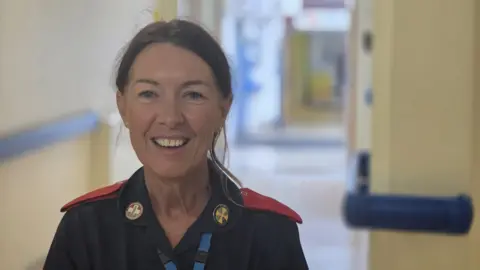 A woman wearing nursing uniform. She has brown hair which is tied back and is smiling at the camera. She is stood in a corridor which is blurred. There are cream walls. 