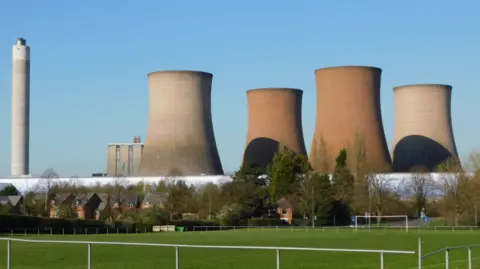 Four large towers, a large chimney and another tall building dominte the skyline, with a football field in the foreground