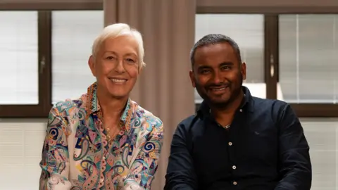 BBC Martina Navratilova, with short white hair, wearing a colourful paisley-print blue and orange collared shirt and several gold chain necklaces. She's sitting next to Amol Rajan, who is wearing a black collared shirt. Both are smiling at the camera. The background is out of focus but there are windows and beige curtains visible.