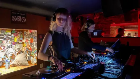 Ellie Tevendale Two women DJ on a set of decks in a dark club.