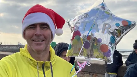 Kate Jennings/BBC The picture shows a person wearing a bright yellow jacket and a red Santa hat, holding a shiny silver star-shaped balloon. The balloon has colourful confetti patterns and text that reads: “YOU’RE A STAR.” The balloon is attached to a pink ribbon. In the background, there is a stone wall, a waterfront area, and several other people dressed in winter clothing. The sky is cloudy with patches of light breaking through.