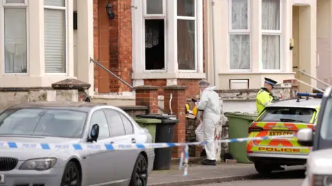 PA Media A terraced house in red brick with white window frames. A person in white overalls is at the entrance to the path leading to the door of the house. A police officer in yellow high-vis is also visible, standing next to a police car. Police tape is stretched across the road.