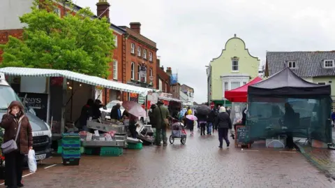 People visiting stalls set up at Fakenham's Market Place on a rainy day. Striped canvas coverings and pop-up marquees are covering the stalls.