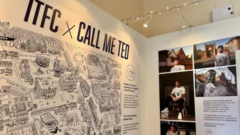 Ipswich Central An exhibition that displays an animated map of Ipswich on a white wall. On the opposite wall are photographs of different people and an information board.