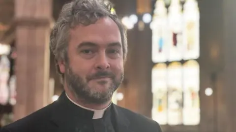 A vicar stood in a church looking into a camera. Stained glass windows can be seen behind him.