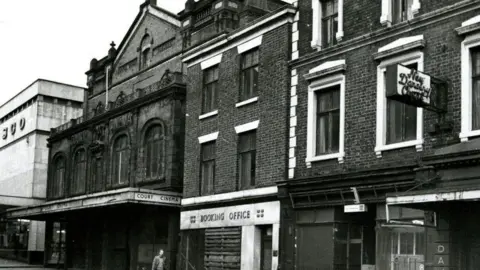A black and white photograph of the front of the theatre building. A white sign with black writing says Court Cinema.