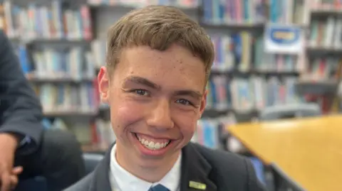 Ethan has short brown hair and is wearing a blue blazer, white shirt and blue and yellow tie. She is sitting in a classroom full of books.