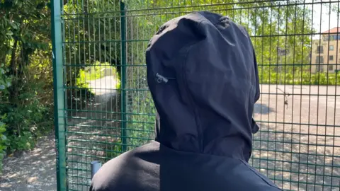 Dylan, who has his hood up, is looking at a playground through a fence