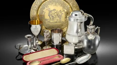 A number of silver items, a golden spoon in a red case, and a golden plate with intricate artwork.