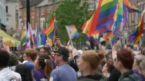 Crowds gather at Pride with many waving rainbow flags in a busy street. The crowds are facing to the left.