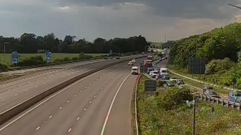 National Highways A grainy CCTV screenshot showing a long line of traffic queuing off a motorway onto the slip road. Beyond the slip road, the motorway is empty.