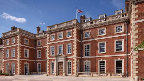 The frontage of an English country house in red brick with ornate brickwork. A flag is flying from a flagpole on the roof against a blue sky. 