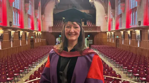 Emma Lucy Cole smiles at the camera and wears graduation robes in the grand hall of the Wills Memorial building in Bristol. It is a grand wood panelled hall with red chairs and lighting