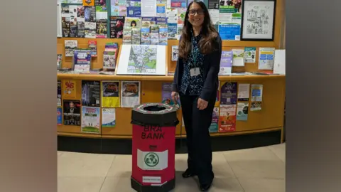 Angela Mathews Angela Mathews, who has long brown hair and glasses, smiles as she stands next to a waist-high red bin with the words "bra bank" written on it. She is standing in front of a display of leaflets and posters.