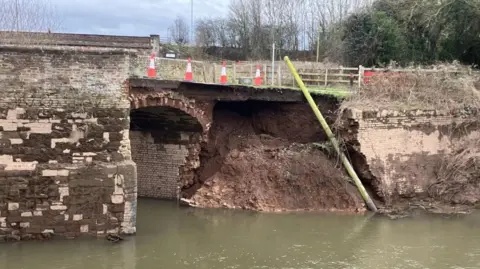 Traffic cones line a section of road across Old Powick Bridge where the supporting wall is completely missing. Bare earth can be seen where the wall has been swept away