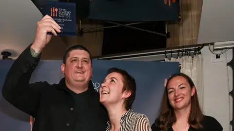 Getty Images Chef Gareth Ward stands on a small stage in front of a “The Good Food Guide” backdrop. He raises an award plaque above his head while smiling, his guest, a woman, looks up at it, smiling, and a woman on the right smiles while holding a microphone and a card.
