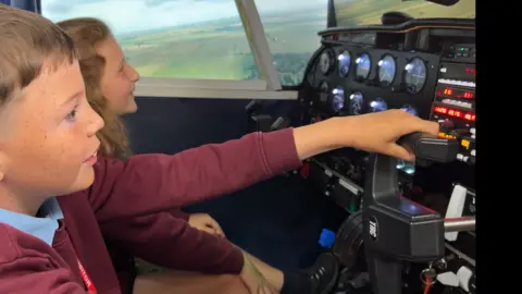 Two primary school pupils, a boy with brown hair and a girl with red hair, in red jumpers inside a flight simulator, you can see the cockpit infront of them and scenery around them