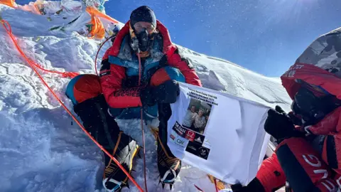 Blue Door Productions/Stan Gaskell/PA Wire Mitch Hutchcraft during his challenge embarking on the "world's longest triathlon". Mr Hutchcraft is sitting on snow. He is dressed in a red puffer suit. He is holding a white banner. The sky is blue.