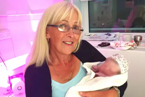 Handout A woman with blond hair and glasses is holding a baby - whose face has been blurred. The woman is wearing a light blue top and black cardigan.