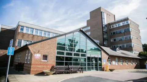 A brick building with a triangle shaped roof and glass window frontage. Two brown benches are outside an entrance. A larger, taller, brown brick building with rows of windows is behind it, to the right. It is a sunny day. 