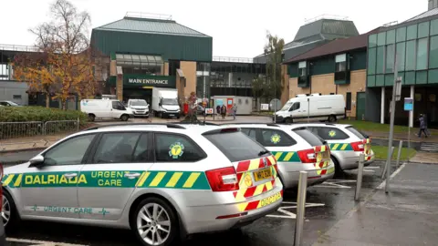 Pacemaker A wide shot of the main entrance to Antrim Area Hospital, a large brown brick building with a green roof. There are several vehicles parked outside the hospital, including three urgent car cars with emergency vehicle markings. 
