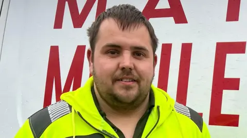 Mousumi Bakshi/BBC Ben Setchell is in the middle stood in front of a white van which has red writing on it. He has short brown hair and beard and is wearing a yellow Hi-Vis jacket. 