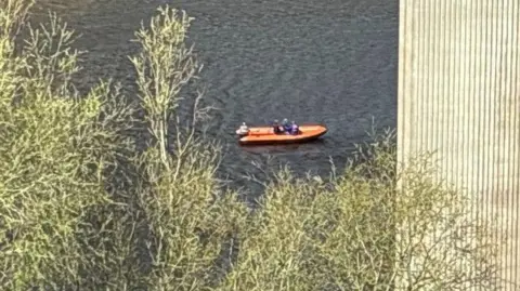 An orange rescue boat on murky water can be seen through the branches of a tree and wooden post. There are three crew members sitting in the vessel.