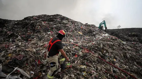 Getty Images A person in red suspenders and a red helmet walks up a gigantic pile of rubbish. An excavator machine is in the background