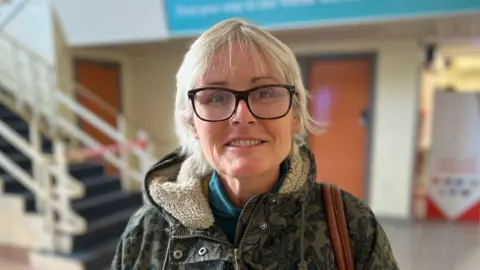 Jennifer Bridge is standing in the airport smiling at the camera. She has short blonde hair and is wearing a pair of glasses. She is wearing a coat with a fleecy hood and a handbag is looped over her shoulder.