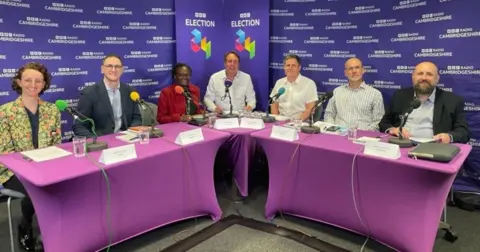 Five men and two women sit round a desk with names on signs in front of them