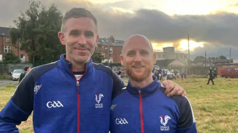 Two French GAA coaches standing on field wearing blue. 