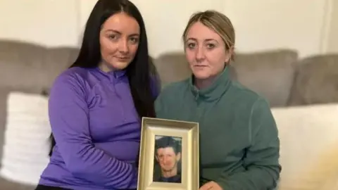 Danielle Bradley is in a purple zipped top and is sitting on a sofa. Danielle has long straight, black hair. Ashling Graham is holding a picture of their father, Jim McFadden. She has short blonde hair.