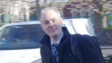 A man with grey hair and a black coat is walking into a court building. This is the defendant James Rouse at a previous court appearance in Portsmouth in January 2025