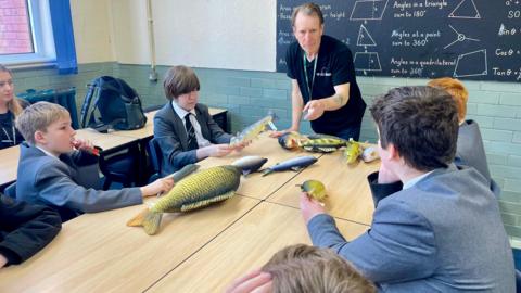 Four pupils sit at a communal desk with soft toy fish in front of them. A teacher stands beside the desk pointing at one of the fish.
