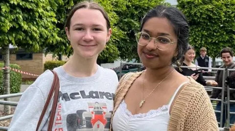 A woman with brown hair tied back is wearing a grey shirt that says 'McLaren'. She is standing next to a woman wearing a white tank top and a knitted cardigan. 